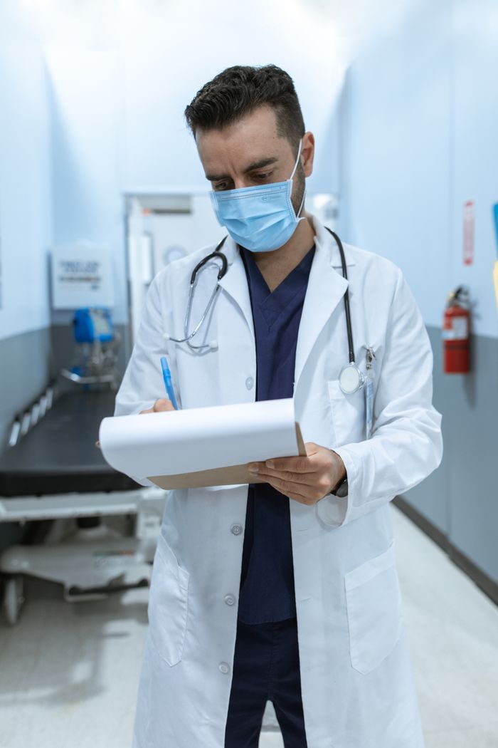Medical professional wearing mask, writing on chart in hospital hallway.